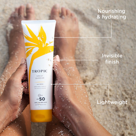 Model holding Tropic Great Barrier Sun Lotion SPF 50 on sandy beach, labelled nourishing & hydrating, invisible finish and lightweight.
